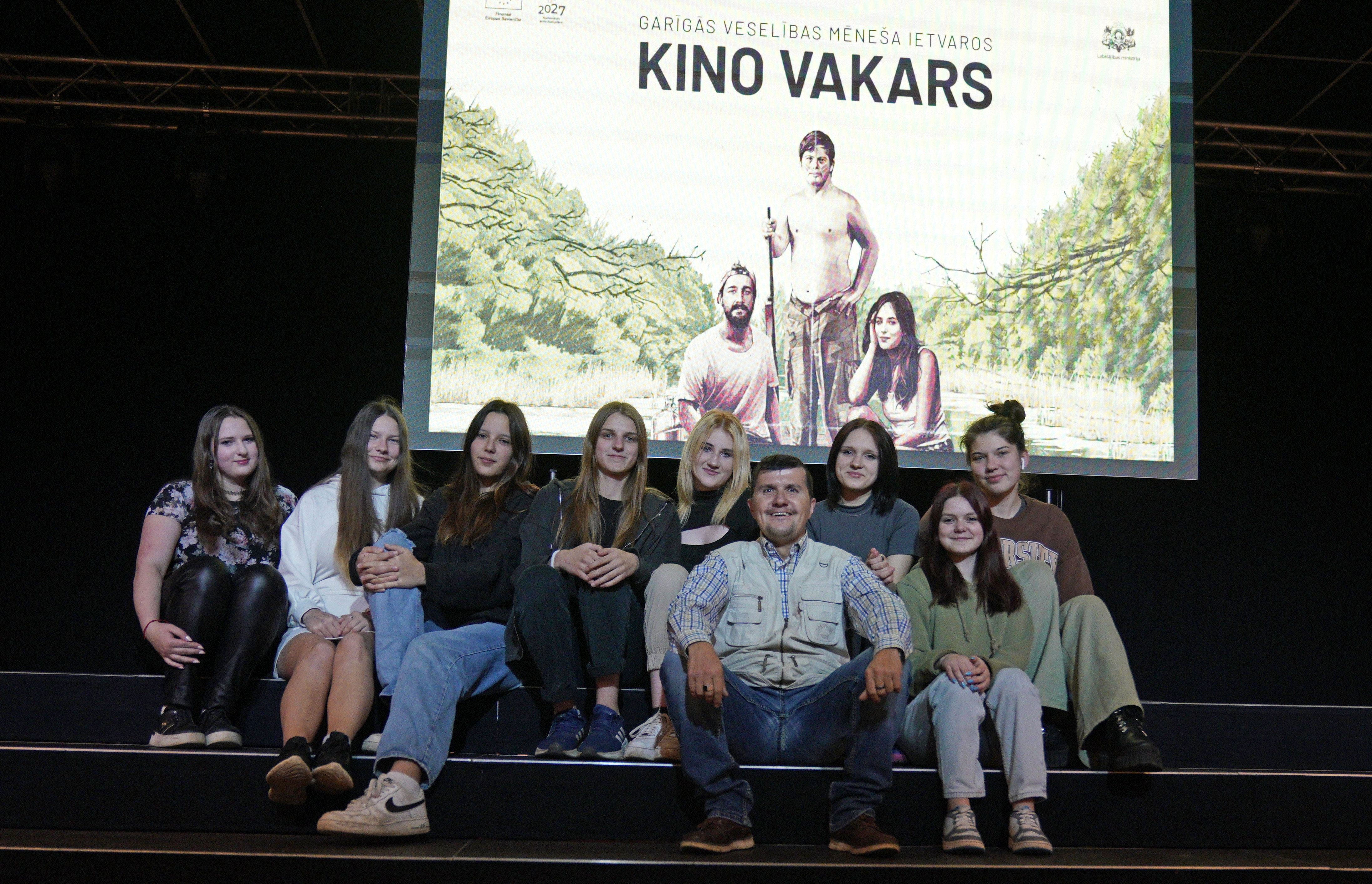 Uz skatuves sēž astoņas skolnieces un vīrietis. Visi smaidot pozē fotogrāfam