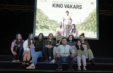 Uz skatuves sēž astoņas skolnieces un vīrietis. Visi smaidot pozē fotogrāfam