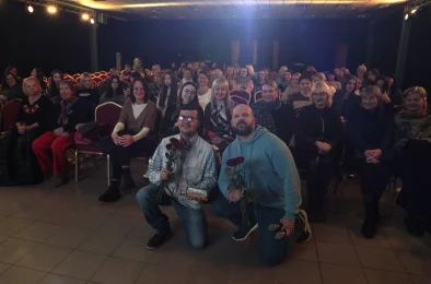 Divi vīrieši notupušies uz viena ceļa ar ziediem rokā. Fonā sēdoša auditorija