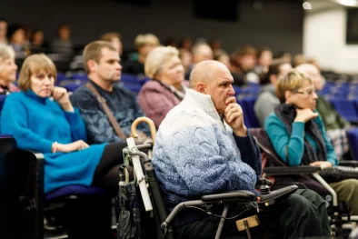 Kino zāle ar sēdošiem cilvēkiem. Priekšplānā kungs gados ratiņkrēslā