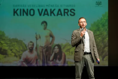 Vīrietis ar mikrofonu rokā uzstājas publikas priekšā