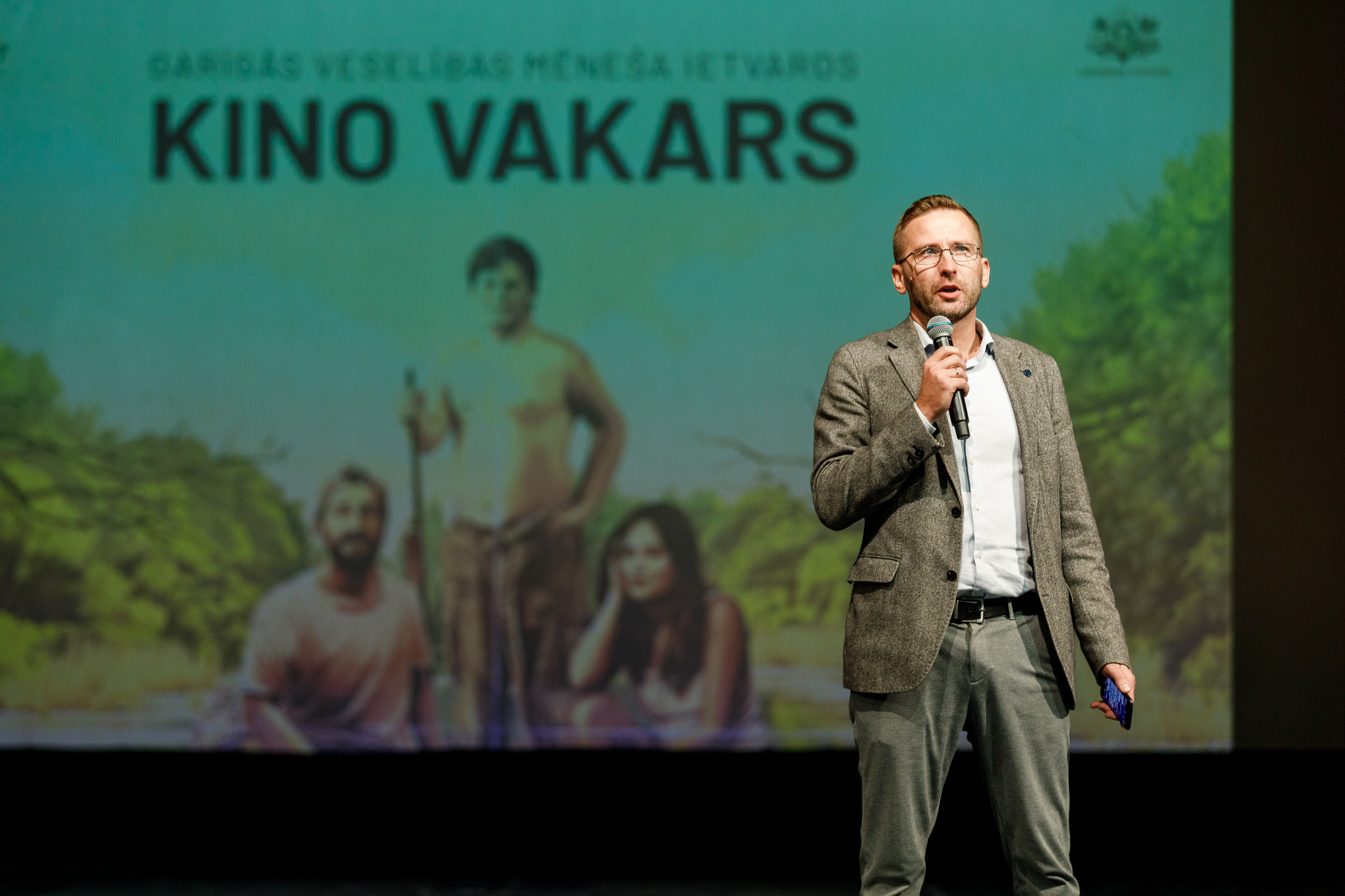 Vīrietis ar mikrofonu rokā uzstājas publikas priekšā