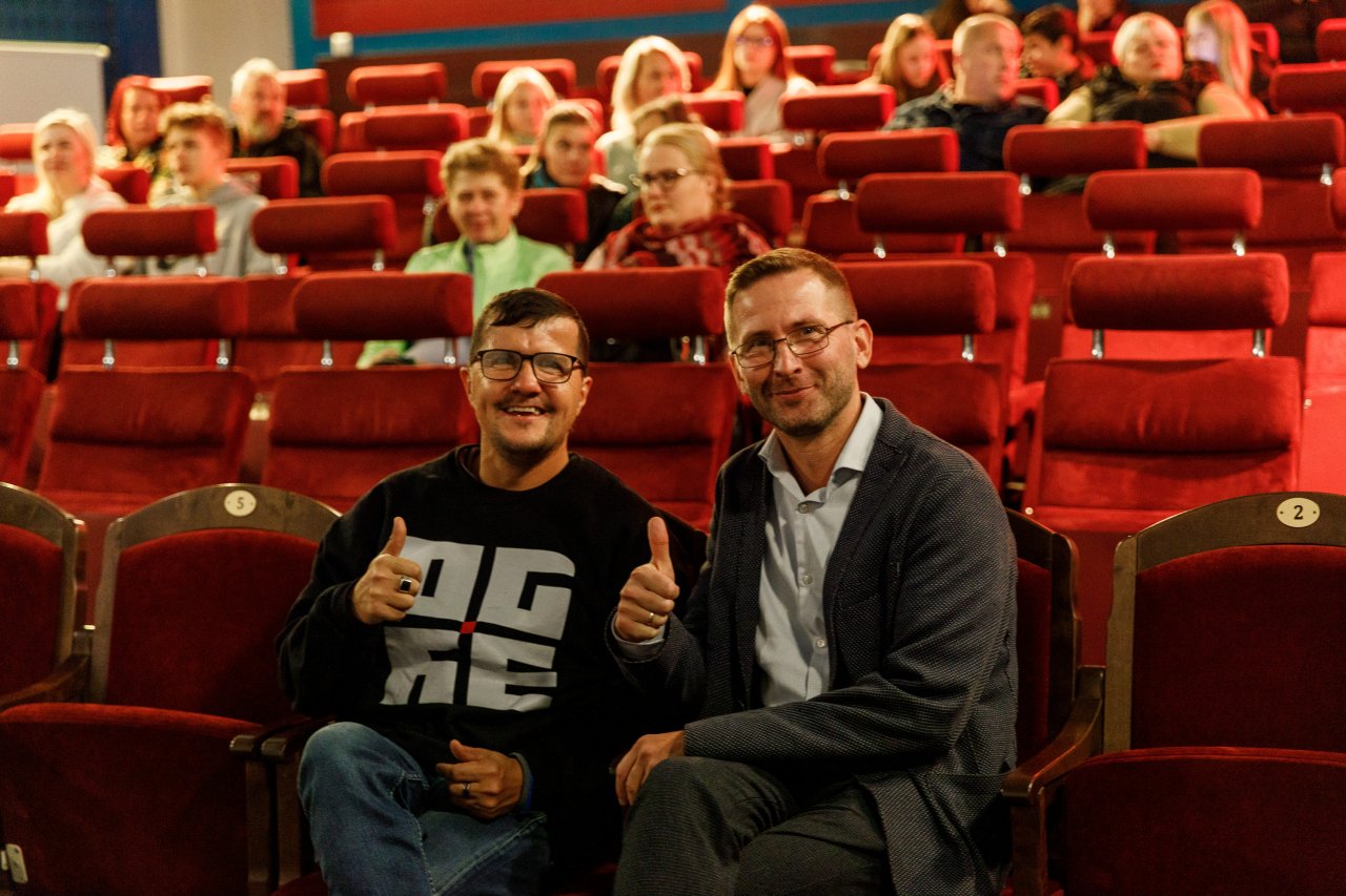 Kino teātris. Sēž divi vīrieši, smaida un rāda paceltus gaisā īkšķus