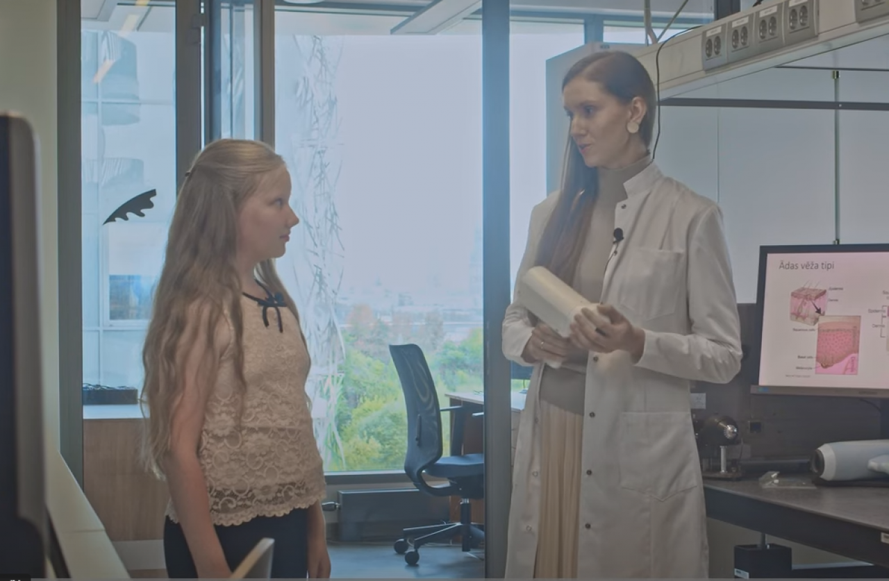 Saite uz iedvesmas stāstu par vēža diagnostiku. Zinātniece stāv ar meiteni laboratorijā, rāda vēža diagnostikas iekārtu